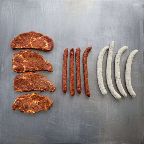 Probierpaket Grillmix S mit mariniertem Schweinehals, Pfefferbeißern und Gourmet-Würsten auf Edelstahlfläche.