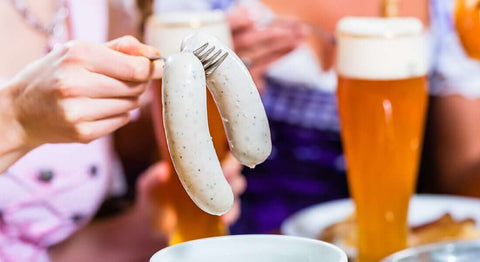 Traditionelle Weißwurst wird mit der Gabel aus dem Sud gehoben, daneben ein Glas Weißbier – typische bayerische Brotzeit