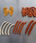 Probierpaket Grillmix mit Gourmet-Würsten, Chili-Krainern, Halsgratsteaks, Hähnchenbruststeaks und Cabanossy, übersichtlich auf Edelstahl angerichtet.