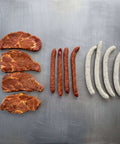 Probierpaket Grillmix S mit mariniertem Schweinehals, Pfefferbeißern und Gourmet-Würsten auf Edelstahlfläche.