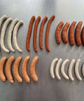 Grillwürste M mit Gourmet-Würsten, Käse-Grillern, Chili-Krainern, Schweinswürsteln und Currywürsten auf Edelstahlfläche.