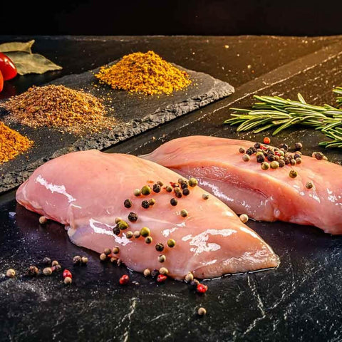 Zwei rohe Hähnchenbrustfilets mit Pfefferkörnern auf dunkler Steinplatte, im Hintergrund Gewürze und Rosmarin