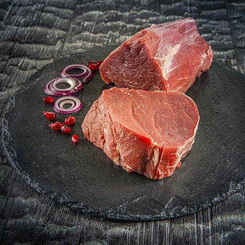 Zwei Stücke rohes Rinderfilet auf einer runden Schieferplatte mit roten Zwiebelringen und Granatapfelkernen