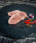 Zwei rohe Schweinefilets auf runder Schieferplatte mit roten Chilis, Pfefferkörnern und Zwiebelringen